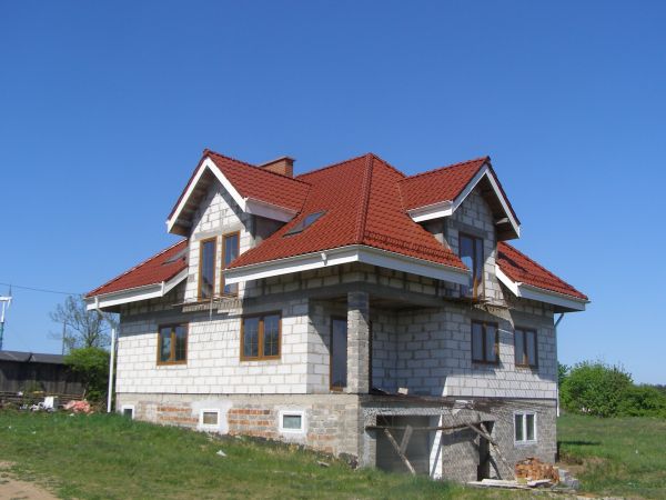 Dom w trakcie budowy z nowym, czerwonym pokryciem dachowym i białymi rynnami, widoczne bloczki betonowe i drewniane podpory.