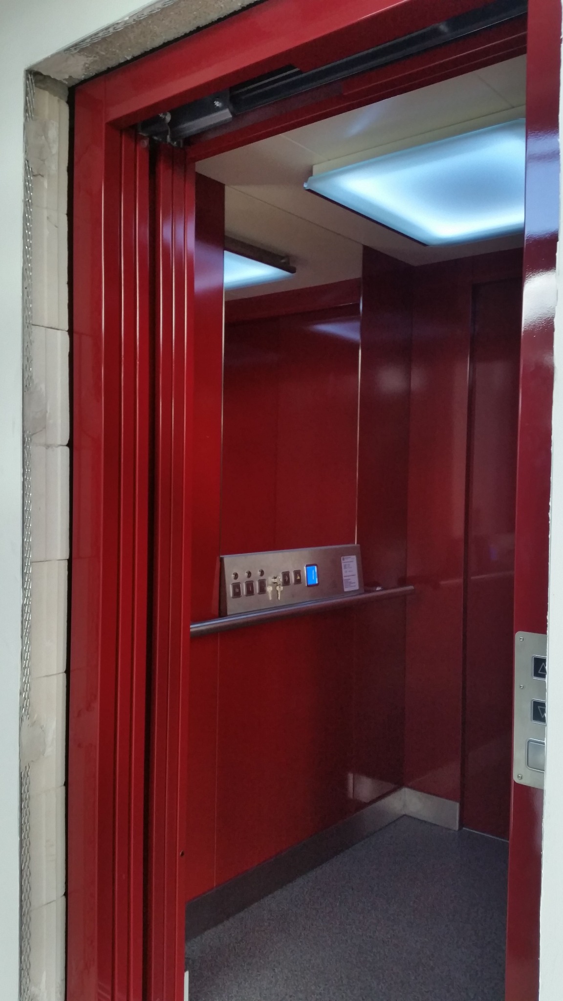 Czerwona kabina windy z częściowo otwartymi harmonijkowymi drzwiami, widoczne panele sterowania i oświetlenie sufitowe.