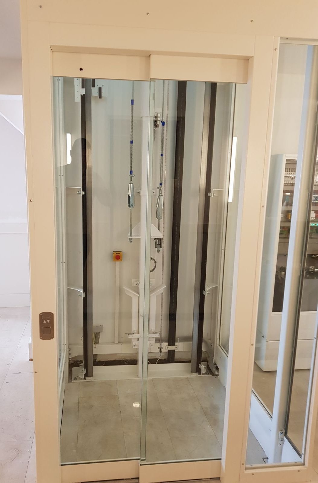 Wnętrze przeszklonej kabiny platformy pionowej z widocznymi elementami konstrukcyjnymi, szynami, linami i przyciskiem awaryjnym.