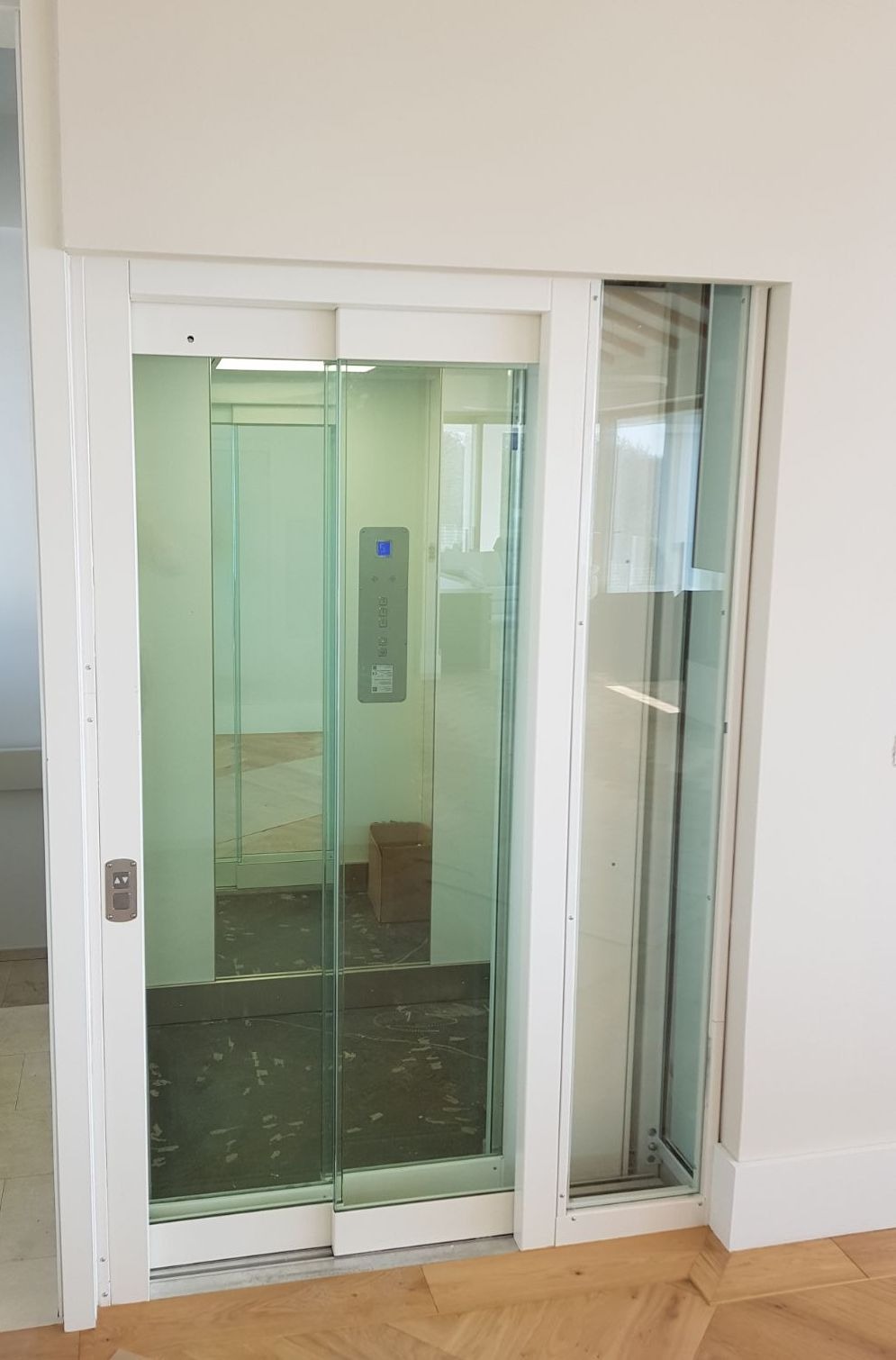 Szklana kabina windy z przesuwnymi drzwiami w jasnym pomieszczeniu, widoczny panel sterowania z niebieskim wyświetlaczem i karton wewnątrz.