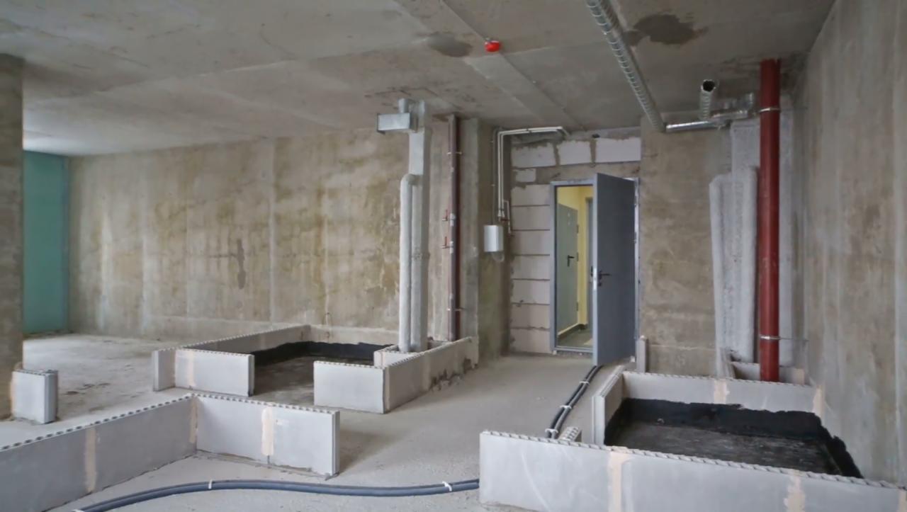 Surowy stan wnętrza z betonowymi ścianami, instalacjami pod sufitem i przygotowanymi wnękami w podłodze, zabezpieczonymi czarną folią, prawdopodobnie pod wylewkę.