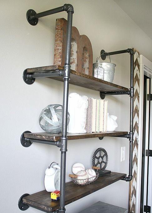 Regał ścienny z rur hydraulicznych i drewnianych półek, dekorowany metalową literą M, ocynkowanym wiaderkiem, książkami, kulą dekoracyjną, zegarem i koszykiem z piłkami do baseballu.