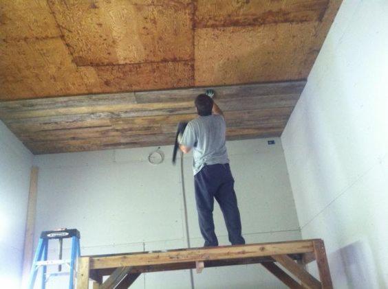 Pracownik na drewnianym rusztowaniu montuje panele drewniane na suficie w pomieszczeniu z surowymi ścianami, widoczna instalacja elektryczna i drabina.