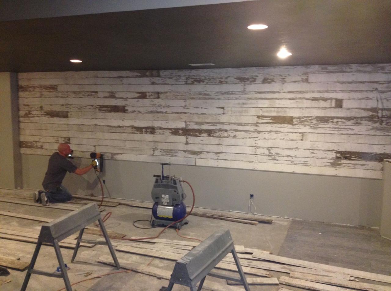 Instalacja dekoracyjnej ściany z desek o efekcie postarzenia: mężczyzna na kolanach przytwierdza deski do ściany za pomocą narzędzia pneumatycznego, widoczny kompresor i rozłożone deski na podłodze.