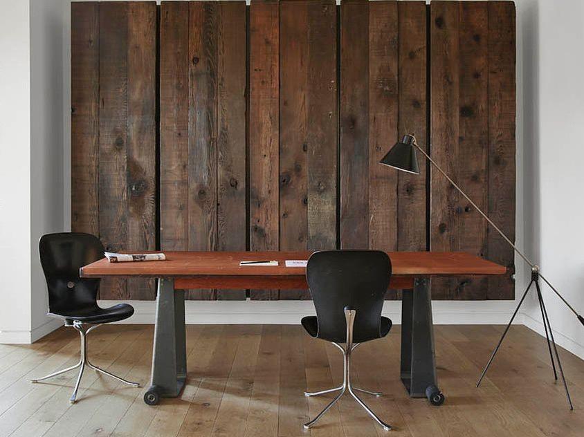 Minimalistyczne biuro z drewnianym panelem ściennym, drewnianym blatem biurka wspartym na metalowych nogach z kółkami, dwoma czarnymi krzesłami i lampą podłogową.