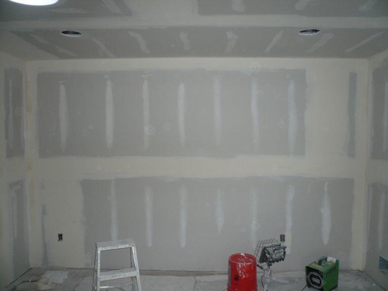 Pokój w trakcie szpachlowania ścian płytami gipsowo-kartonowymi, widoczna aluminiowa drabina, czerwone wiadro i nagrzewnica w tle.