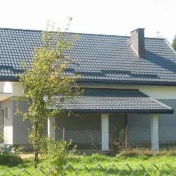 Nowoczesny dom jednorodzinny z szarą dachówką karpiówką, jasnoszarą elewacją i brązowym kominem, widok zza ogrodzenia z zielenią.