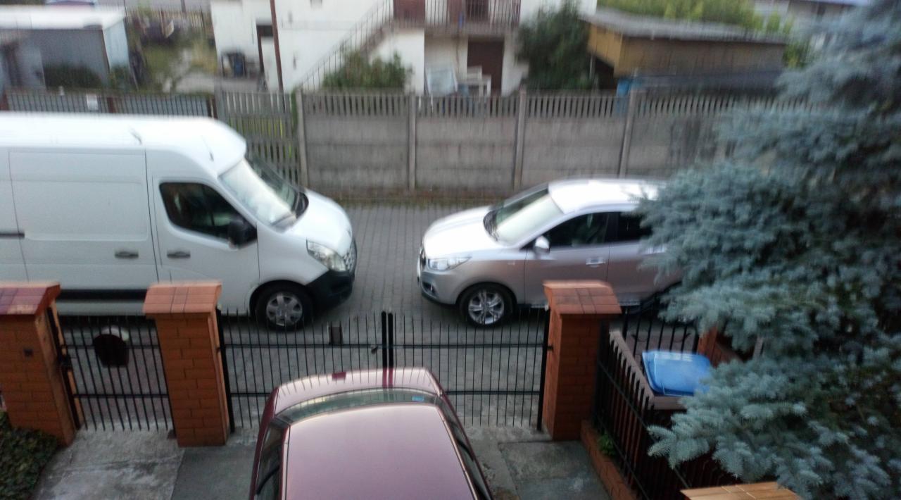 Widok z góry na zaparkowany biały samochód dostawczy obok srebrnego samochodu osobowego, oddzielone czarnym, kutym ogrodzeniem z ceglanymi słupkami, na tle szarego betonowego ogrodzenia i zabudowań.