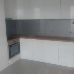 Minimalistyczna kuchnia z białymi, lakierowanymi frontami szafek, drewnianym blatem i szarym panelem szklanym między szafkami górnymi i dolnymi, widoczny piekarnik do zabudowy.