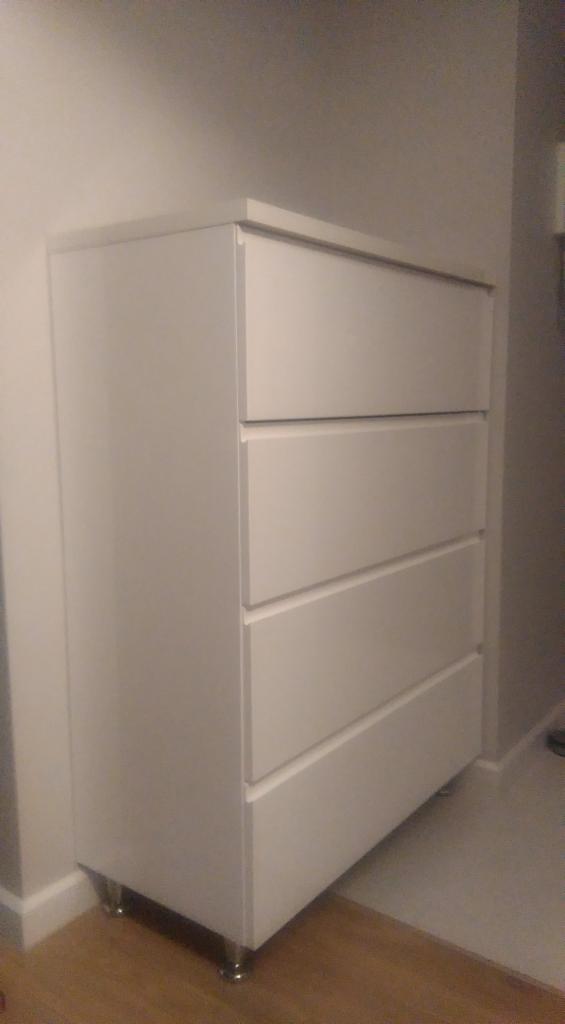 Biała komoda z czterema szufladami na metalowych nóżkach, ustawiona przy szarej ścianie w pomieszczeniu z drewnianą podłogą.