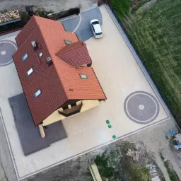 Widok z góry na dom z beżową elewacją i czerwoną dachówką, otoczony rozległym podjazdem z dekoracyjną nawierzchnią z okrągłymi wzorami, ogrodzeniem i trawnikiem.
