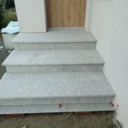 Trzy stopnie schodowe z jasnoszarego kamienia prowadzące do drzwi wejściowych, widoczne czerwone podkładki poziomujące i cegły pod spodem, świeżo ułożone.