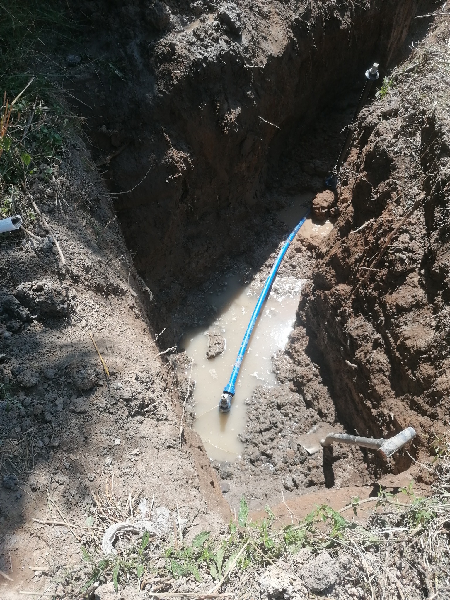 Wykop z wodą, w którym widoczna jest niebieska rura z przyłączem, obok leży szpadel.