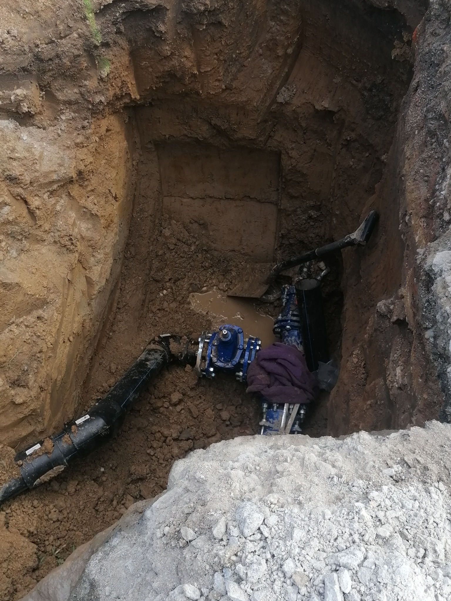 Wykop z widocznym przyłączem wodno-kanalizacyjnym z metalowymi elementami i czarną rurą, częściowo zalany wodą, otoczony ziemią i gruzem.