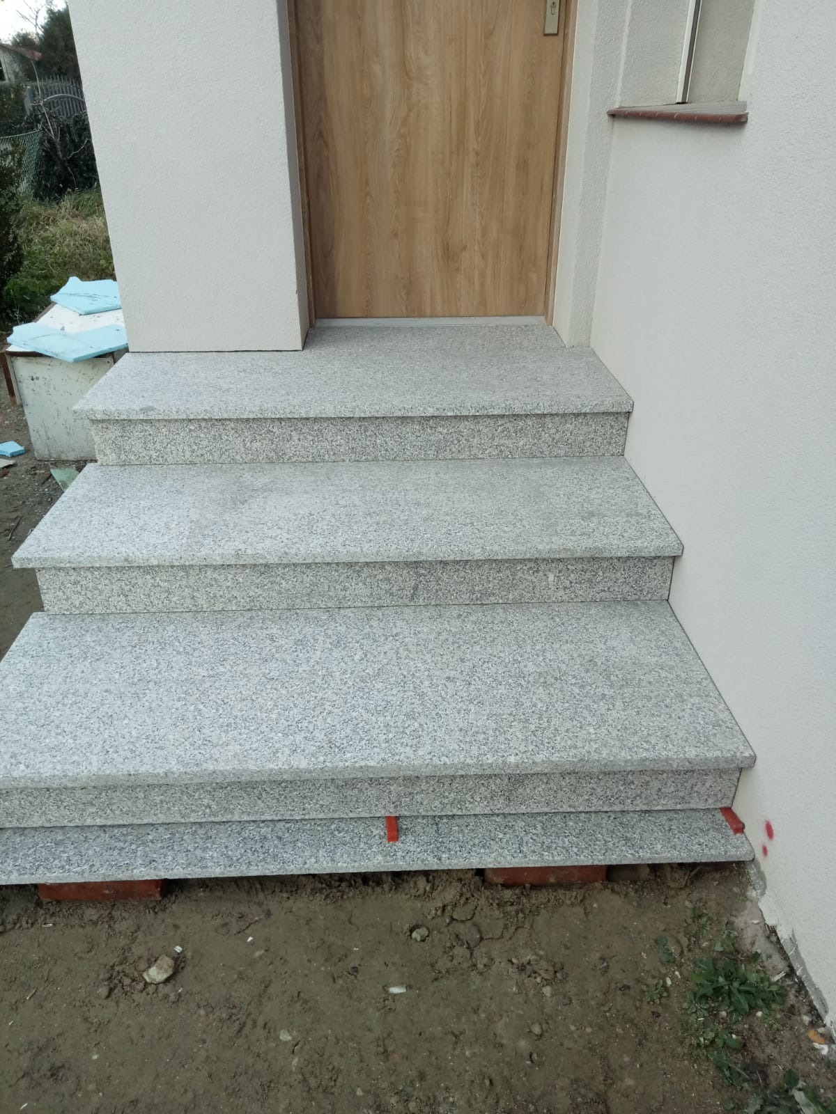 Trzy stopnie schodowe z jasnoszarego kamienia prowadzące do drzwi wejściowych, widoczne czerwone podkładki poziomujące i cegły pod spodem, świeżo ułożone.