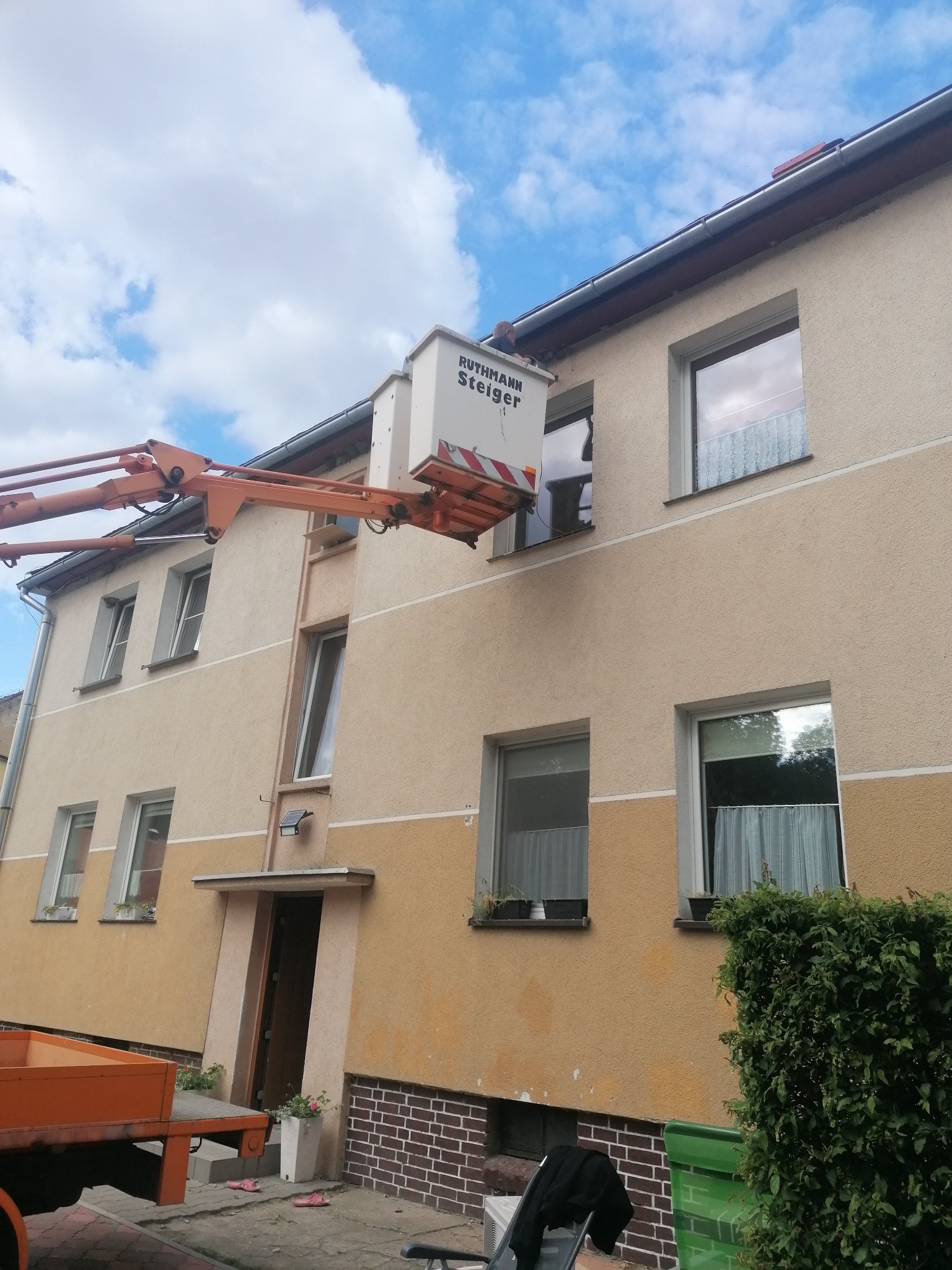Prace na wysokościach przy budynku mieszkalnym wykonywane z podnośnika koszowego marki Ruthmann Steiger, widoczne okna z firankami i rośliny w doniczkach.