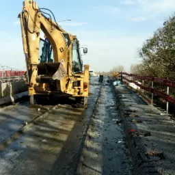 Żółta koparka na moście w trakcie frezowania nawierzchni, widoczne ślady frezowania i barierki ochronne.