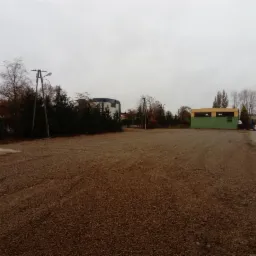 Przygotowany teren pod budowę, wyrównana powierzchnia z drobnym żwirem, w tle budynek magazynowy i drzewa w jesiennej scenerii pod pochmurnym niebem.