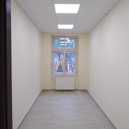 Baza Kolorów - Puste, świeżo odnowione pomieszczenie z beżowymi ścianami, jasną podłogą imitującą drewno i oknem z kratami, widok zza otwartych drzwi.