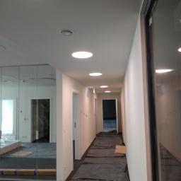 Baza Kolorów - Korytarz biurowy w trakcie przygotowań do prac wykończeniowych, podłoga zabezpieczona folią ochronną, ściany świeżo pomalowane na biało, widoczne okrągłe lampy sufitowe.