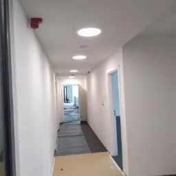 Korytarz biurowy w trakcie przygotowań do malowania: podłoga zabezpieczona folią i kartonem, świeżo pomalowane ściany na biało, okrągłe lampy sufitowe, czujnik dymu.