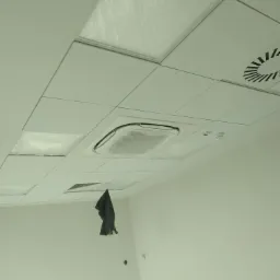 Biały sufit podwieszany z kwadratowymi panelami, niektóre zabezpieczone folią, widoczny klimatyzator kasetonowy w folii ochronnej i kratka wentylacyjna, fragment ściany z widocznymi puszkami...