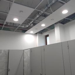 Baza Kolorów - Wnętrze biurowe w trakcie wykańczania, widoczne kabiny sanitarne, białe ściany i sufit podwieszany z oświetleniem, instalacje pod sufitem.