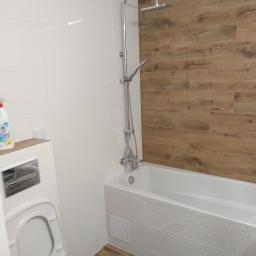 Baza Kolorów - Nowoczesna łazienka z białymi i drewnopodobnymi płytkami, kabina prysznicowa z deszczownicą, umywalka z toaletą w zabudowie, widoczny płyn do czyszczenia na blacie.