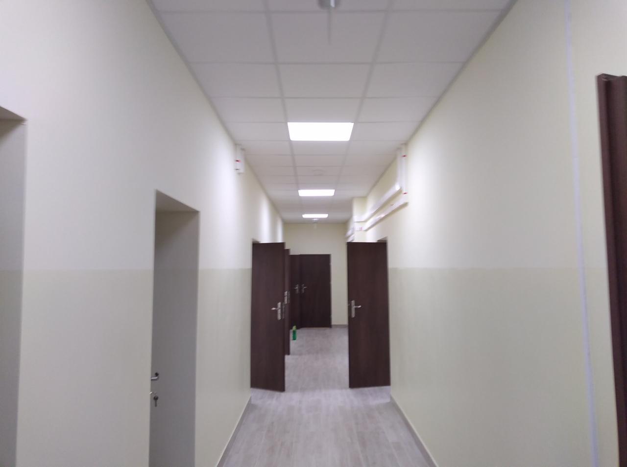 Długi korytarz biurowy z kremowymi ścianami, ciemnymi drzwiami i podwieszanym sufitem z kwadratowymi panelami oświetleniowymi, widok w głąb z otwartymi drzwiami.