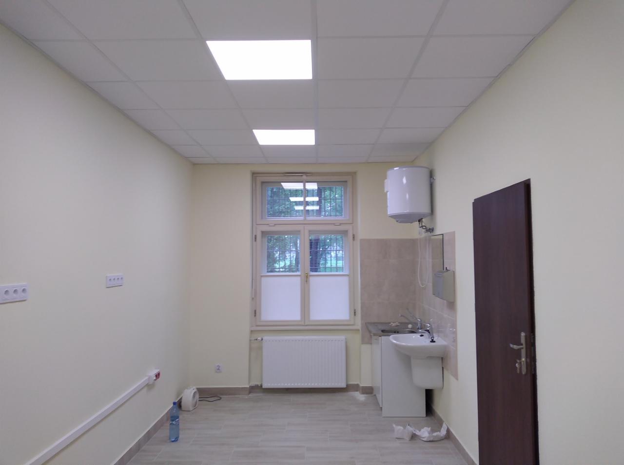 Jasne pomieszczenie z umywalką i bojlerem, ściany pomalowane na kremowo, na podłodze płytki imitujące drewno, okno z roletami.