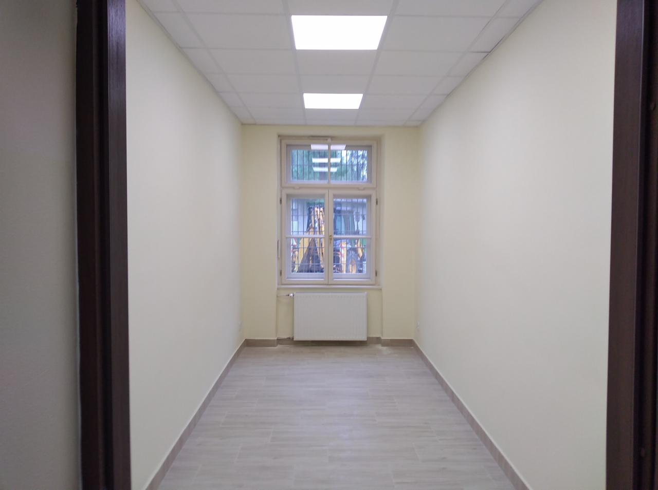 Puste, świeżo odnowione pomieszczenie z beżowymi ścianami, jasną podłogą imitującą drewno i oknem z kratami, widok zza otwartych drzwi.