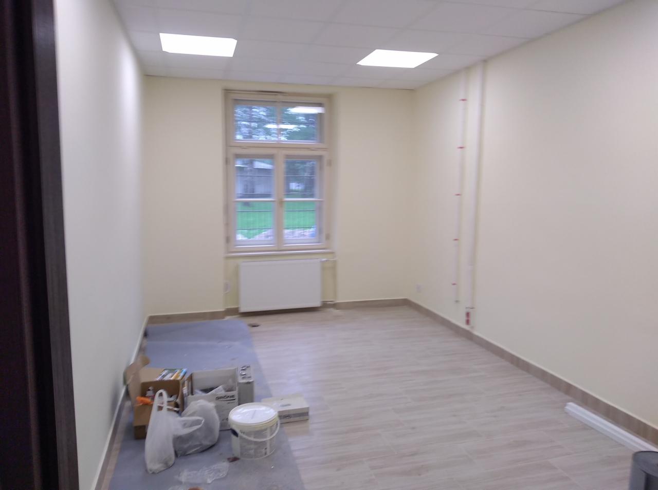 Pokój po odświeżeniu koloru ścian na jasny odcień, widoczne akcesoria malarskie na podłodze, okno z grzejnikiem, płytki podłogowe imitujące drewno.