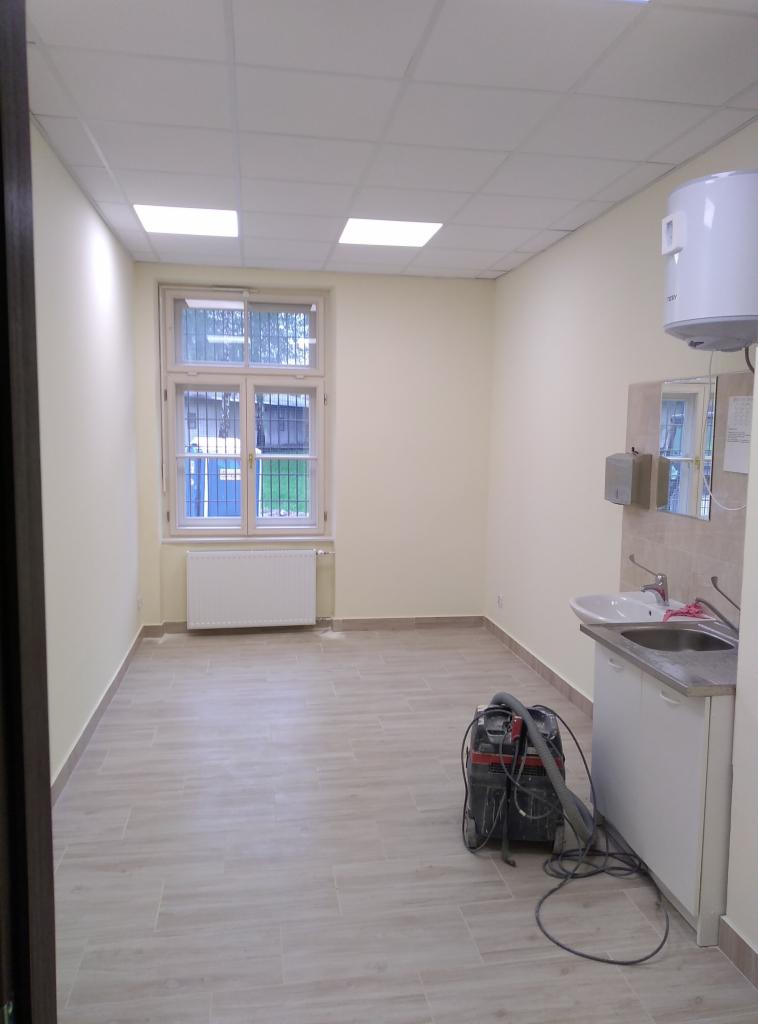 Pomalowane na jasno biuro z oknem, grzejnikiem, umywalką i odkurzaczem na podłodze z płytek imitujących drewno, gotowe do użytku.