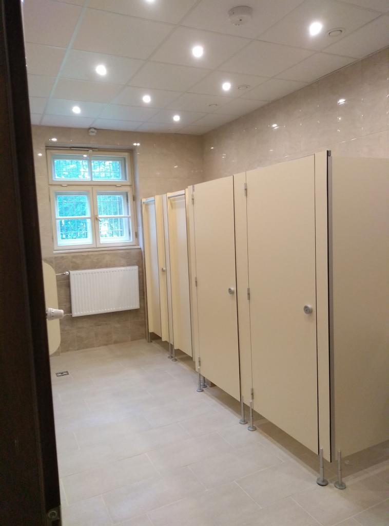 Wykończona toaleta publiczna w Krakowie z kabinami sanitarnymi w beżowej kolorystyce, jasnymi płytkami na ścianach i podłodze oraz oknem z widokiem na zieleń.