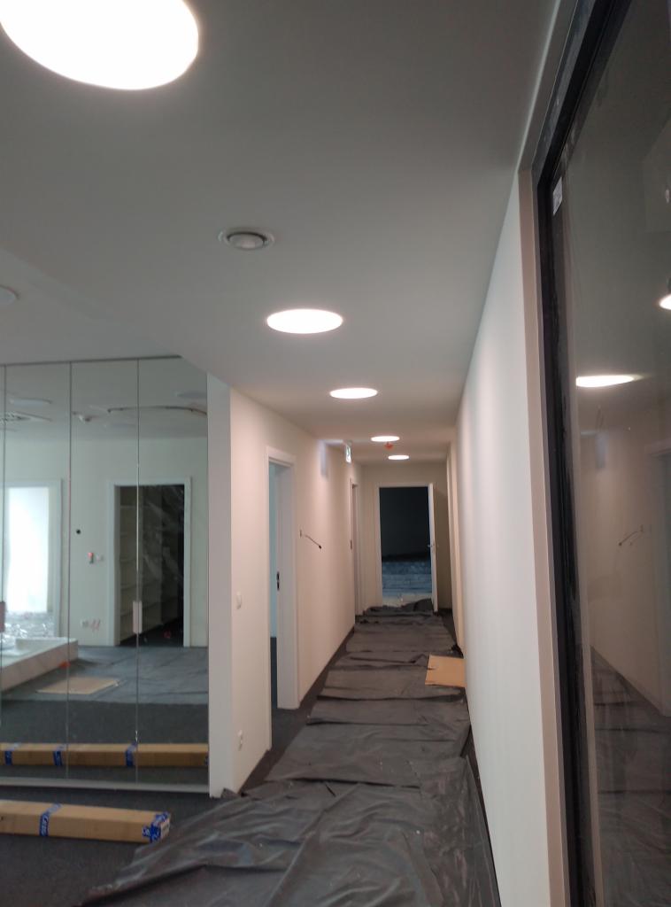 Korytarz biurowy w trakcie przygotowań do prac wykończeniowych, podłoga zabezpieczona folią ochronną, ściany świeżo pomalowane na biało, widoczne okrągłe lampy sufitowe.