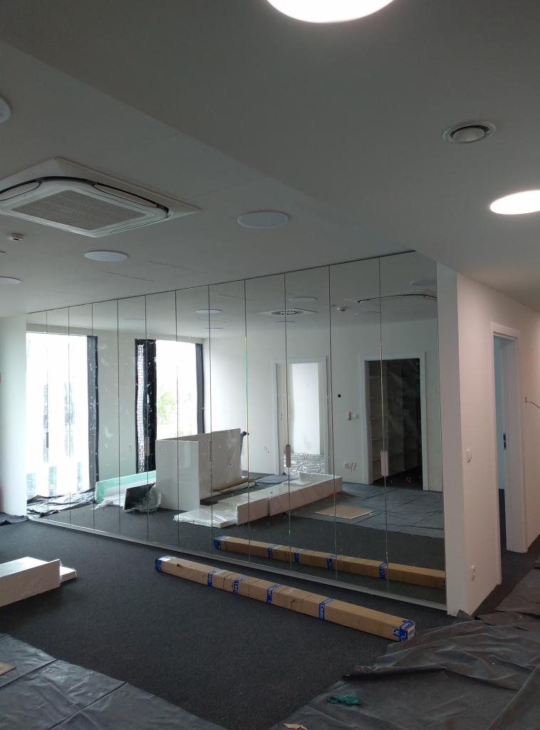 Montaż lustrzanej ściany z segmentów w biurze, widoczne kartony z materiałami, folia ochronna na podłodze i elementy wyposażenia w tle.