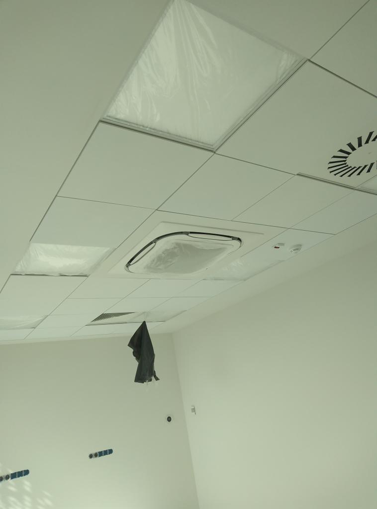 Biały sufit podwieszany z kwadratowymi panelami, niektóre zabezpieczone folią, widoczny klimatyzator kasetonowy w folii ochronnej i kratka wentylacyjna, fragment ściany z widocznymi puszkami...
