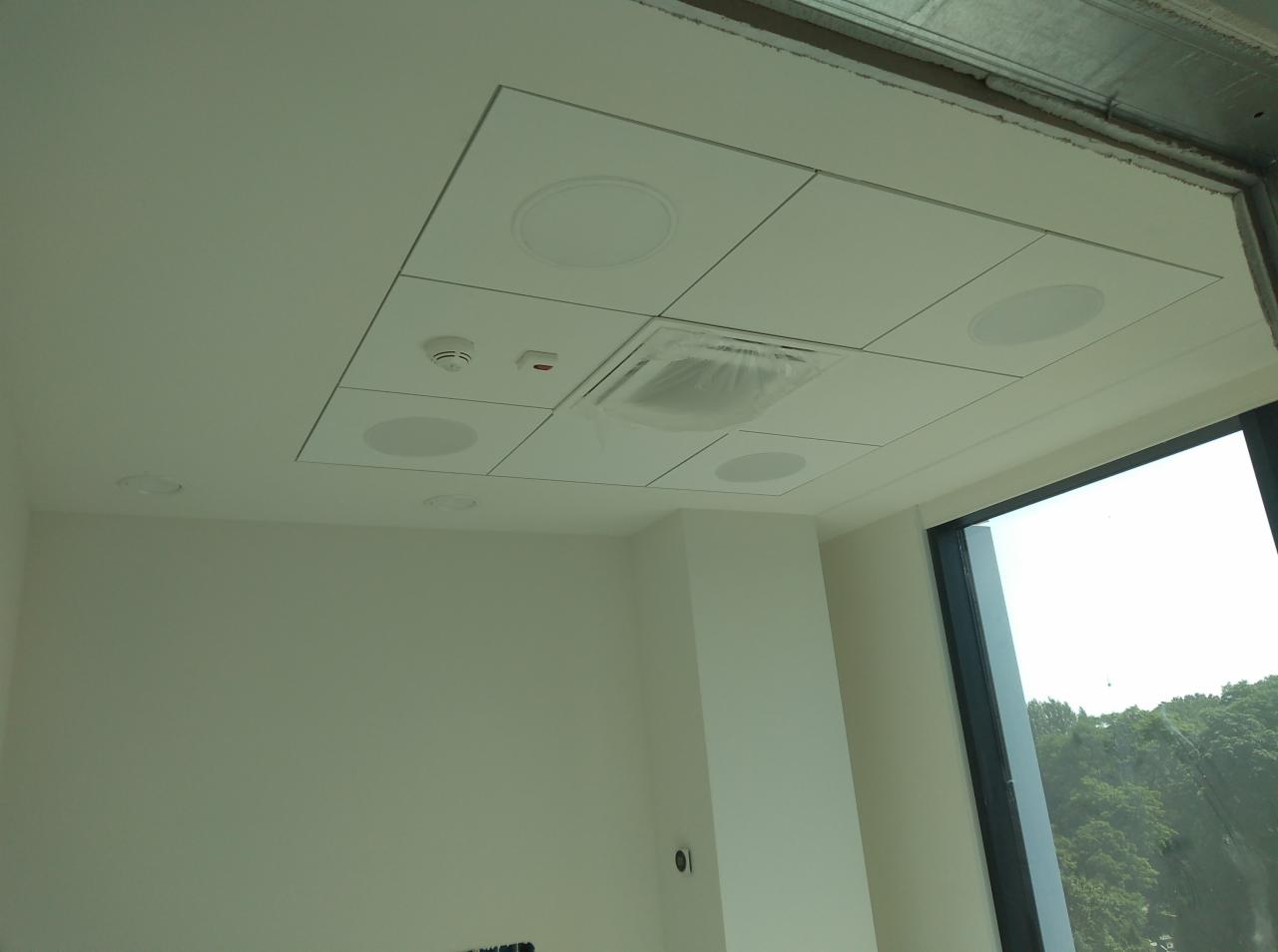 Biały sufit kasetonowy z wbudowanymi głośnikami i kratką wentylacyjną zabezpieczoną folią, widok na jasne wnętrze biura z dużym oknem.
