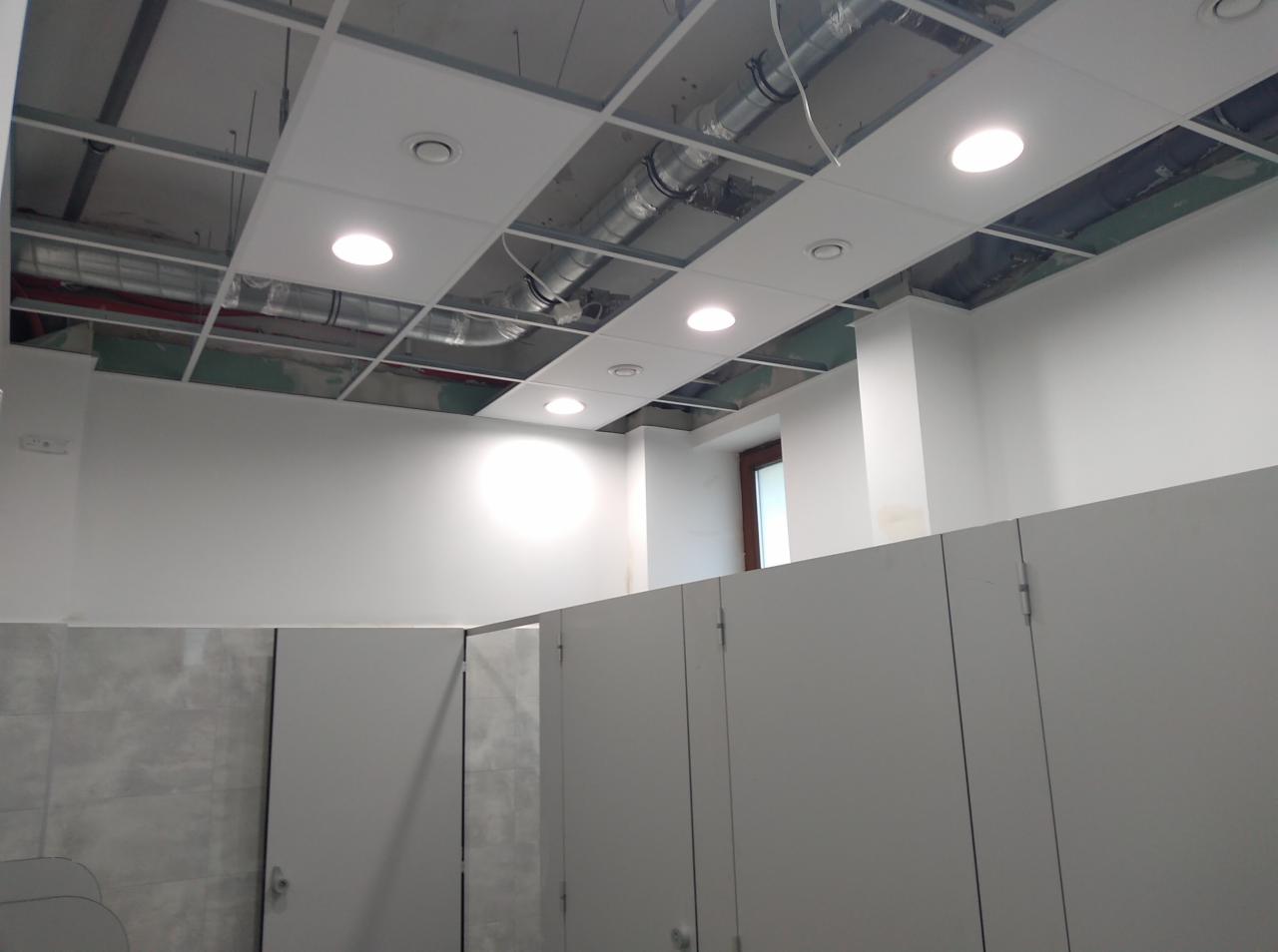Wnętrze biurowe w trakcie wykańczania, widoczne kabiny sanitarne, białe ściany i sufit podwieszany z oświetleniem, instalacje pod sufitem.