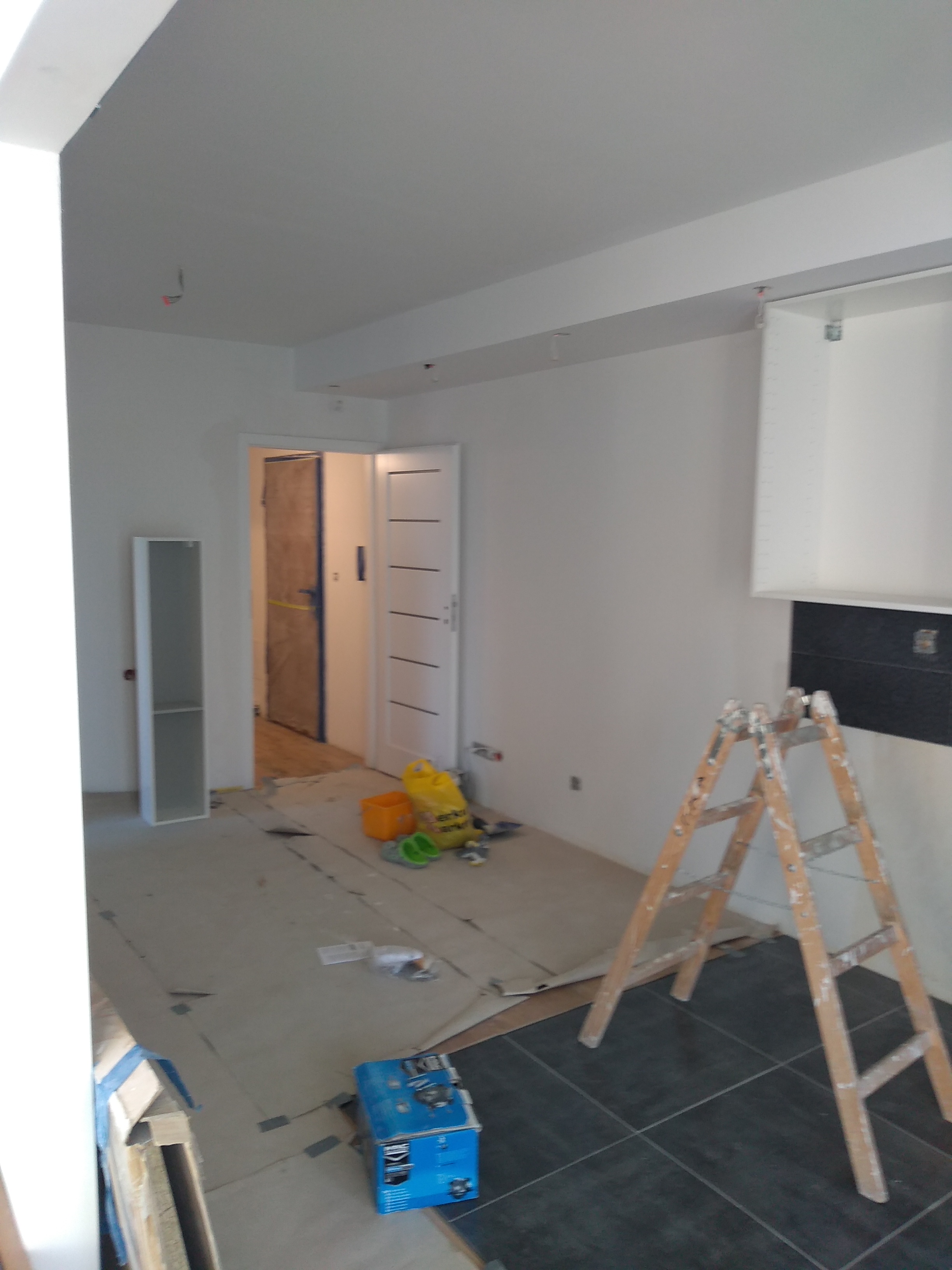 Wnętrze remontowanego mieszkania z białą ścianą, drewnianą drabiną opartą o ścianę z ciemnymi płytkami, podłogą zabezpieczoną kartonem, żółtym workiem z materiałami budowlanymi i otwartymi białymi...