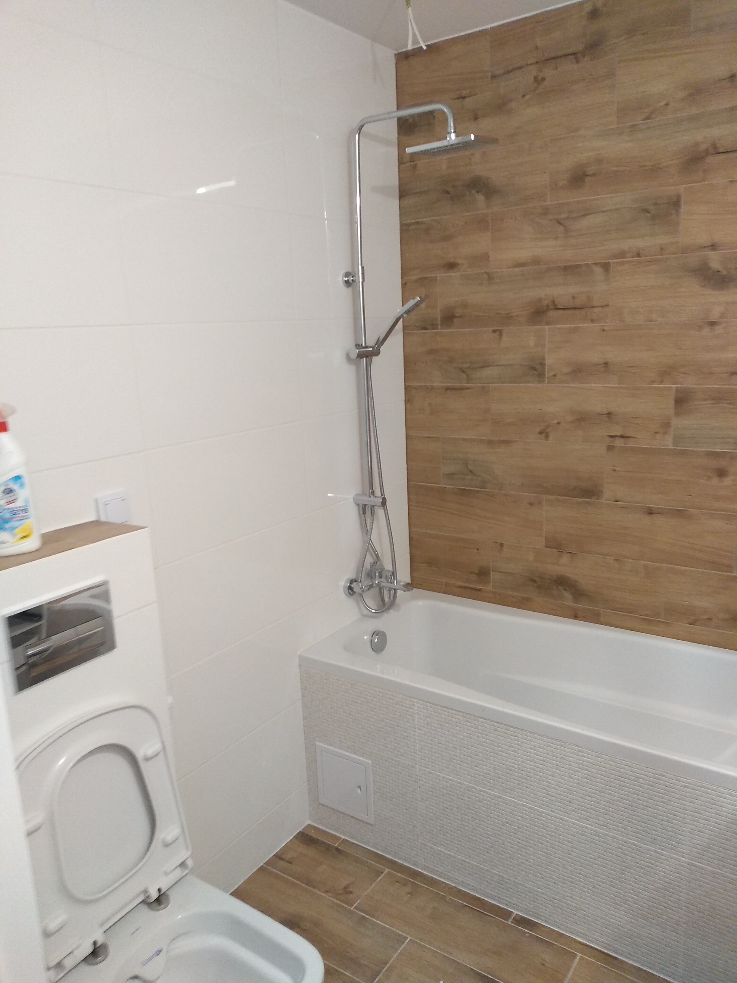 Nowoczesna łazienka z białymi i drewnopodobnymi płytkami, kabina prysznicowa z deszczownicą, umywalka z toaletą w zabudowie, widoczny płyn do czyszczenia na blacie.