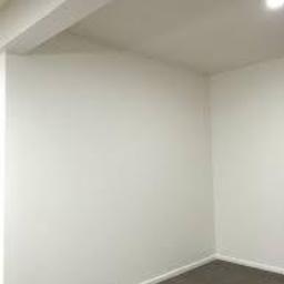 Jeremi Kluczek - Pusty, pomalowany na biało pokój z szarą wykładziną podłogową. Widoczne narożniki ścian, sufit z widoczną instalacją i oświetlenie punktowe.