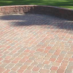 Jeremi Kluczek - Ułożona kostka brukowa w odcieniach czerwieni i szarości tworząca okrągły taras, ograniczony niskim, zakrzywionym murem z cegły, z trawnikiem w tle.