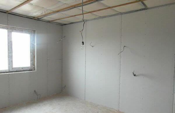 Pokój w trakcie wykańczania: ściany wyłożone płytami gipsowo-kartonowymi, widoczne przewody elektryczne, surowy sufit z izolacją i metalową konstrukcją, okno z PCV.