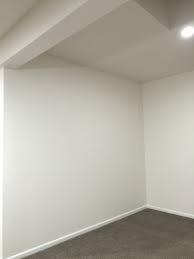 Pusty, pomalowany na biało pokój z szarą wykładziną podłogową. Widoczne narożniki ścian, sufit z widoczną instalacją i oświetlenie punktowe.