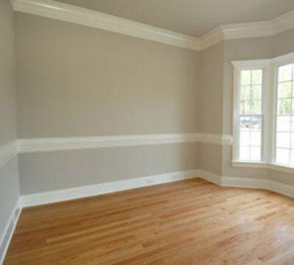 Pusty pokój po remoncie z parkietem w odcieniach brązu, szare ściany z białymi listwami dekoracyjnymi i wykuszem okiennym.