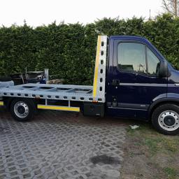 Auto serwis oponex - Niebieski bus z platformą lawety bez ładunku, widok z boku na tle zielonego żywopłotu i kostki brukowej.