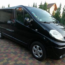 Auto serwis oponex - Czarny bus Renault Trafic z przyciemnianymi szybami, zaparkowany na kostce brukowej przed ogrodzeniem z tujami i domem w tle.