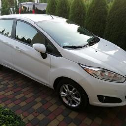 Auto serwis oponex - Biały Ford Fiesta hatchback zaparkowany na kolorowej kostce brukowej, tło stanowią zielone tuje i ciemny samochód dostawczy.