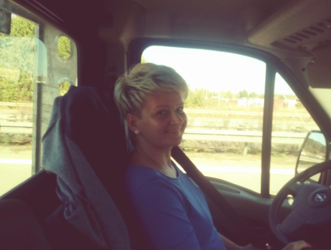 Uśmiechnięta blond kobieta w niebieskiej bluzce siedzi za kierownicą pojazdu dostawczego, widok na otoczenie przez okno.